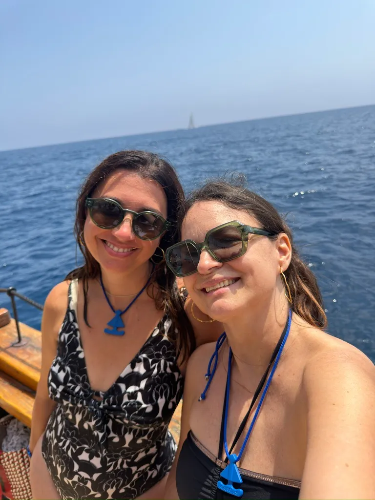 Dos mujeres sonriendo en la cubierta de un velero con el mar azul detrás y colgantes de ancla azul