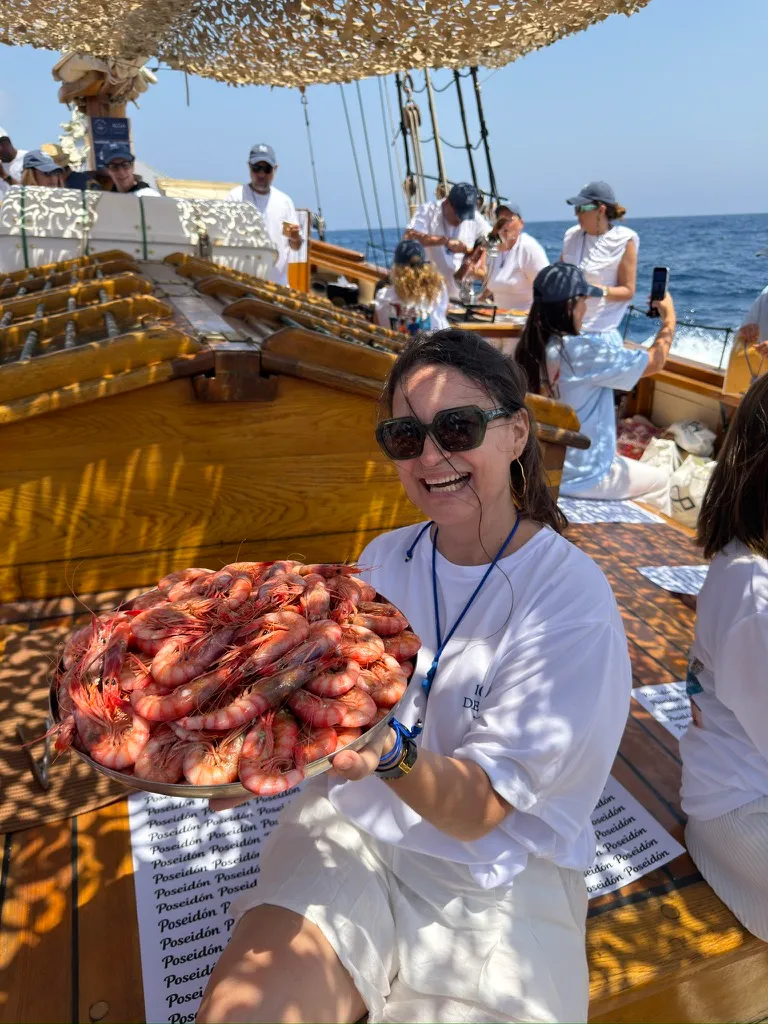 Mujer con gafas de sol sonriendo con una bandeja de gambas rojas en la cubierta de madera de un velero, con el mar detrás