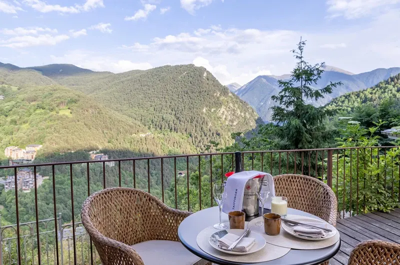 Mesa de terraza con sillas de mimbre en Jou la Borda, con el valle verde y las montañas de fondo