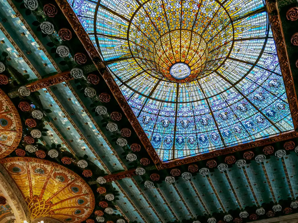 Cúpula de vitral de colores del Palau de la Música Catalana vista desde abajo, rodeada de rosas de cerámica