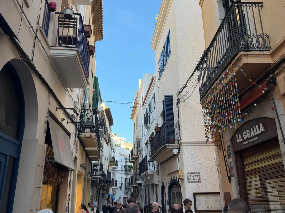 Calle estrecha del casco antiguo de Sitges con fachadas blancas, balcones de hierro y gente paseando