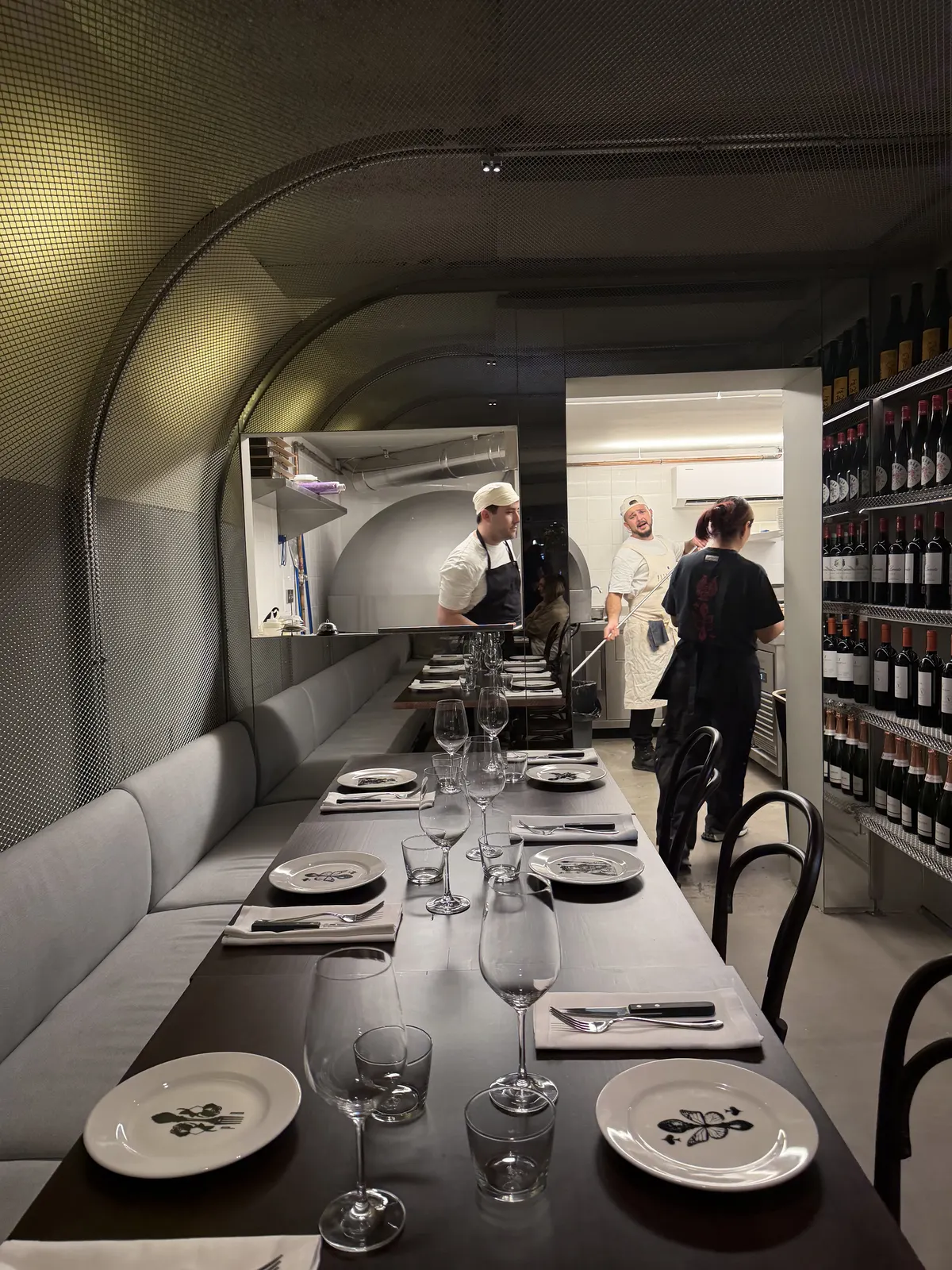 Mesa larga de Pizzabar con platos y copas, el horno de pizza al fondo y el equipo trabajando en la cocina abierta